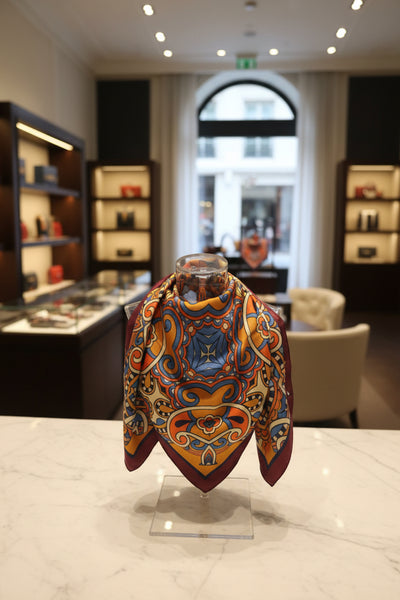 A square scarf laid flat on a white table, featuring a vibrant mandala and paisley pattern in yellow, blue, orange, and cream, with a deep maroon border. A lime green wall is visible in the background.
