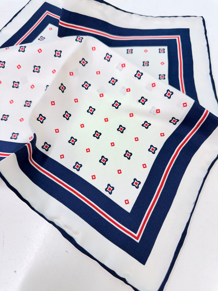 A square white silk scarf with a navy blue border and a repeating pattern of small red and navy geometric motifs, laid flat on a white surface.