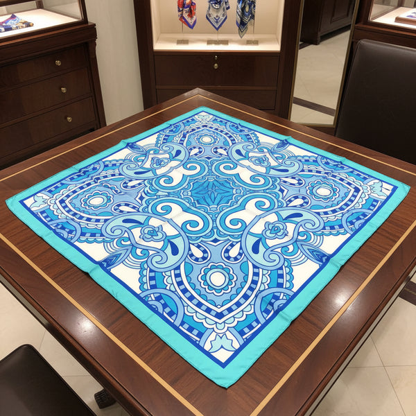 A square silk scarf with an intricate blue and white mandala pattern and a turquoise border, laid flat on a white surface.
